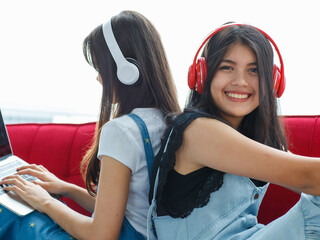 Close-up portrait shot of a cute smiling young Thai-Turkish teenagers sitting back to back on a red couch and listening to music. Mixed-race sisters living together with one playing a laptop