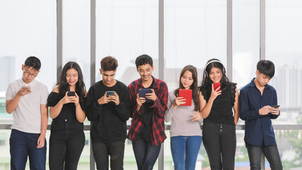 Group of seven young male and female teenagers standing and playing their smartphones and tablet. Junior friends using a mobile phone without talking. Concept of social media addicted in adolescent