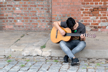 Fototapeta premium young latin boy enjoying playing the guitar while sitting on the floor with a brick wall background, underground, urban