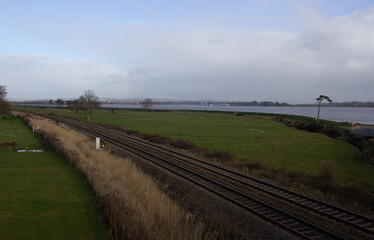 double track railway line in typical English farm land with a large river in the background with ablue sky and white clouds