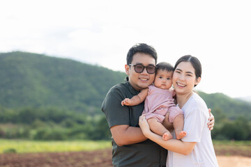 Obraz premium Happy asian family father mother child daughter on nature background . Parents hug little baby in arm standing outdoor with sunny day.