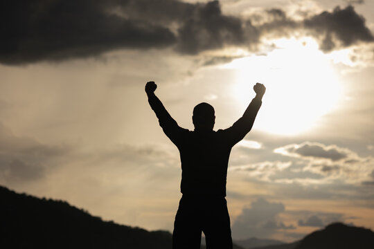 Silhouette Young man raised hand in the air with success goal on sunset background. Man standing on top of the mountain freedom life concept.