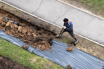 A worker digging a ditch along a pavement in summer.