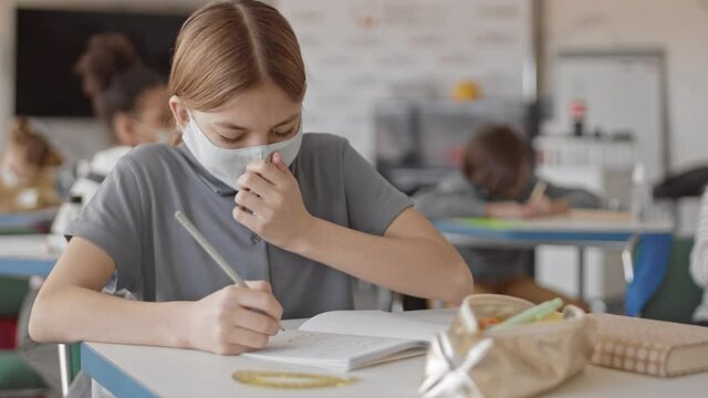 PAN medium slowmo of teenage schoolgirl in face mask sneezing while her classmates disinfecting stationery objects with sanitizer in blurred background sitting together in modern class during pandemic