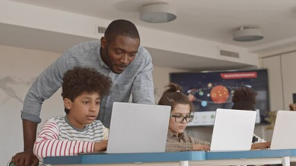 Medium slowmo of male Afro-American school teacher looking at laptop screen controlling work of his talented Mixed-race student of 5 grade at IT lesson - Powered by Adobe