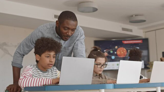 Medium slowmo of male Afro-American school teacher looking at laptop screen controlling work of his talented Mixed-race student of 5 grade at IT lesson