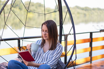 Female sitting in cozy egg hanging swing chair near lake and holding mag and reading a book....