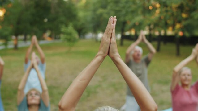 Close up tilt down shot of young woman holding Namaste and talking to group of senior people while leading outdoor yoga class in park