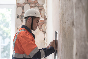 A man in a helmet and a work uniform aligns the walls with a spatula and putty. Copy space	