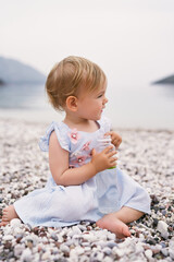 Little girl in a dress sits on a pebble beach and holds a bottle of water in her hands