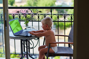 Caucasian toddler boy in diaper plays on laptop on terrace on sunny day. Chromackey screen laptop....