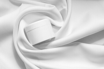 Top view of white jar for packaging cosmetic products, in soft tissue folds. Close-up, monochrome...