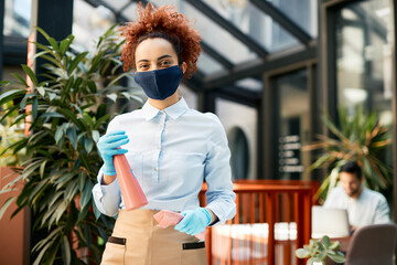 Smiling waitress holding disinfection spay bottle while working in a cafe during coronavirus...