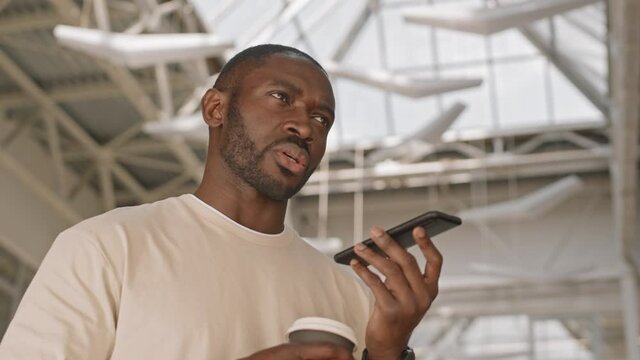 Low-angle waist-up shot of young bearded handsome African-American man in white T-shirt making business phone call talking in speakerphone sipping coffee indoors