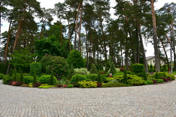 A view of a big, well maintained miniature garden full of shrubs, herbs, trees, and other kinds of flora located next to a circular pavement or pathway seen near the forest in Poland