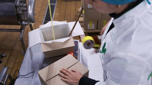 A factory worker manually packs a filled cardboard box on a conveyor belt