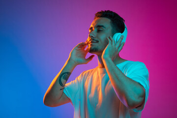 Portrait of Latina young man in white headphones listening to music with closed eyes isolated on gradient purple pink background in neon light.