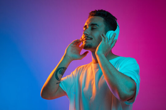 Portrait Of Latina Young Man In White Headphones Listening To Music With Closed Eyes Isolated On Gradient Purple Pink Background In Neon Light.