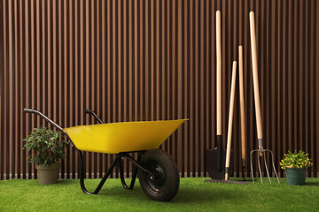 Wheelbarrow, gardening tools and plants on green grass near wood slat wall - obrazy, fototapety, plakaty