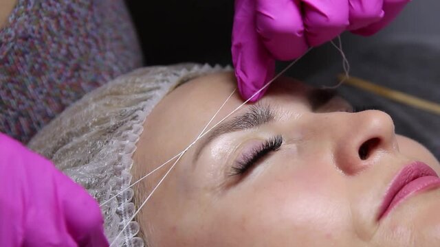performing professional procedures for the correction and shaping of women's eyebrows in a beauty salon. Close-up of the client's face and the hands of the stylist working with a special thread to out