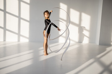 Little girl practising rhythmic gymnastics with a gymnastic tape at white sunny dance room. Wide view with copy space and shadows on the background
