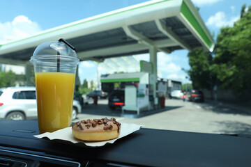 Plastic cup of juice and doughnut on car dashboard at gas station. Space for text