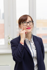 young stylish pretty business woman in a blue jacket and glasses by the window in the office at her workplace is satisfied with the work done