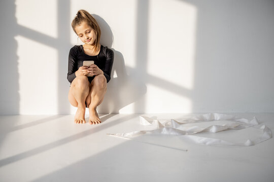 Little girl sitting with smart phone during a break in rhythmic gymnastics class on a white wall background. Female zoomer or generation z in phone during sports