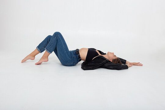 portrait of young caucasian attractive woman with long brown hair in blue jeans and black leather jacket on white background. skinny pretty lady lying on floor at studio with bare feet