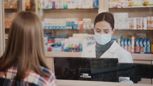 A serious professional pharmacist woman wearing a protective mask is serving the client with his order standing at the cashbox