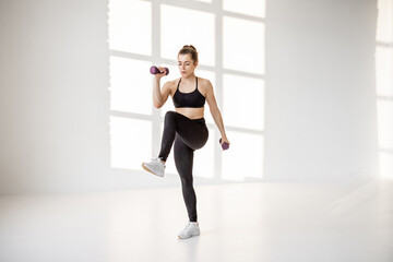 Fototapeta premium Beautiful and confident athletic woman doing cardio with dumbbells at white room. Training fitness or aerobics alone
