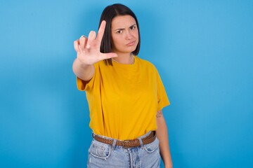 young beautiful brunette girl with short hair standing against blue background making fun of people with fingers on forehead doing loser gesture mocking and insulting.