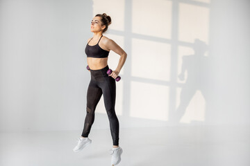Obraz premium Beautiful and confident athletic woman doing cardio with dumbbells at white room. Training fitness or aerobics alone