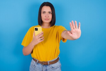 young beautiful brunette girl with short hair standing against blue background using and texting with smartphone with open hand doing stop sign with serious and confident expression, defense gesture