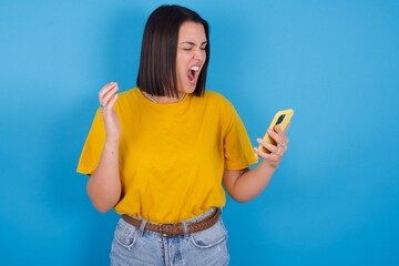 Angry young beautiful brunette girl with short hair standing against blue background screaming on the phone, having an argument with an employee. Troubles at work.