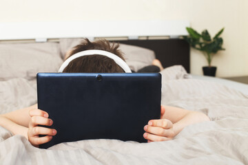 child boy lying on bed and covered his face with a pc tablet with wireless headphones