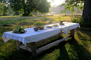 Fototapeta na wymiar Outdoors summer scene party table on sunset. Old wooden table under trees with food plate. Midsummer celebrating in Latvia, Ligo festive