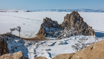 A picturesque two-headed mountain devoid of vegetation rises above the frozen Lake Baikal. Cracks on steep slopes. A motor road on the snowy ice. Clear blue sky. Mount Shamanka.