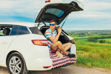 young male traveler sitting in the trunk of his car, man having a rest during a trip, outdoors with...