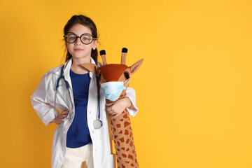 Little girl with eyeglasses, stethoscope dressed as doctor hugging toy giraffe on yellow...