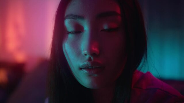 Close up face young beautiful asian woman look at camera sitting on sofa at neon light. Alone, room, evening, couch, single, pretty, attractive. Slow motion