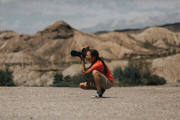 Crouched down woman photographer framing and taking photos in Tabernas Desert, Spain, in a sunny day, with an orange color t-shirt.