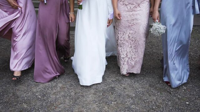 Girls in long dresses go forward. The bride in a white dress and girlfriends in satin dresses walk on the gravel in shoes and sandals
