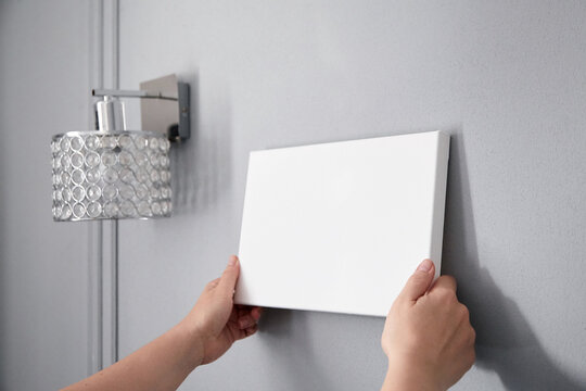 White canvas in female hands with gray wall background. Woman hanging blank picture mockup on wall with sconce