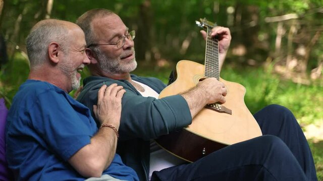 Closeup slow motion of two gay men enjoying nature camping with tent with pride flag in forest on river playing guitar.