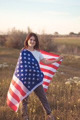 4th of July, Independence day. American freedom concept. Young woman patriot wrapped in the US national flag in a field at sunset. Selective focus