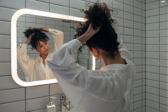 Young mixed race woman looking herself in the mirror in modern washroom. Girl makes a bun in her curly hair. Female making herself a ponytail.