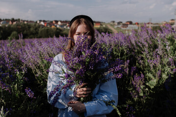 Young woman in shirt holding bouquet of purple flowers covering face standing alone among field in...
