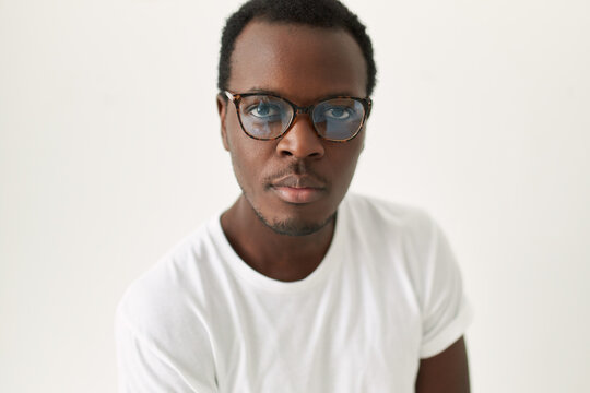 Close up image of handsome young dark skinned man with trimmed stubble posing isolated wearing stylish glasses and white tee looking at camera with confident facial expression. Eyewear and vision