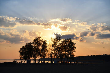 Bright rays of the setting sun. Orange sky and clouds in a hot haze. The beach is filled with vacationers and swimmers. Reflections of the sunset on the water. Trees on the shore. Evening resort.